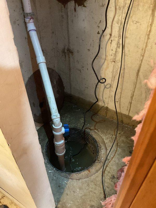 # Sump Pump System Assessment and Commercial Drainage Evaluation

Conducted a comprehensive drain and sewer estimate for a commercial basement utility space. Inspected existing sump pump installation housed in a concrete basin containing standing water, evaluating the operational status of the black submersible pump unit and associated white PVC discharge piping system with multiple connectors and joints. Assessed electrical wiring configuration and power supply connections running to the pump equipment. 

Examined concrete floor conditions throughout the space, noting moisture staining and water infiltration patterns indicative of potential drainage concerns. Evaluated basement waterproofing infrastructure including spray foam insulation application on concrete block walls and exposed pink fiberglass insulation in wall cavities. Documented backup battery system positioned in the utility area for emergency pump operation during power outages. 

Provided detailed assessment findings and