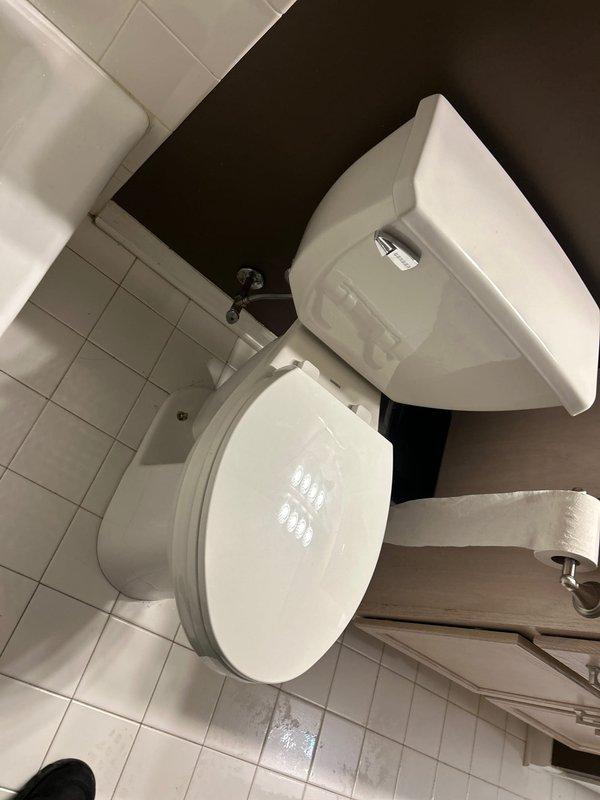 Completed toilet inspection and assessment in residential bathroom. Documented existing conditions of standard two-piece white porcelain toilet installation, including tank, bowl, and flush mechanism. Verified proper placement and functionality of fixture against dark wall with white tile flooring. Confirmed toilet paper holder mounting and overall bathroom plumbing configuration for service records.