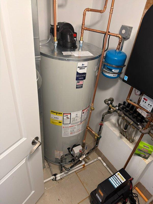 Completed same-day gold-level water heater installation in residential utility space. Installed new Bradford White tanked water heater with complete plumbing connections including copper supply lines, blue-handled isolation valve, expansion tank mounted to wall, and black circulation pump on top of unit. Connected PVC drainage piping at floor level and integrated white plastic manifold distribution system with multiple valve connections. All connections properly secured with appropriate fittings and joints, system tested and operational.