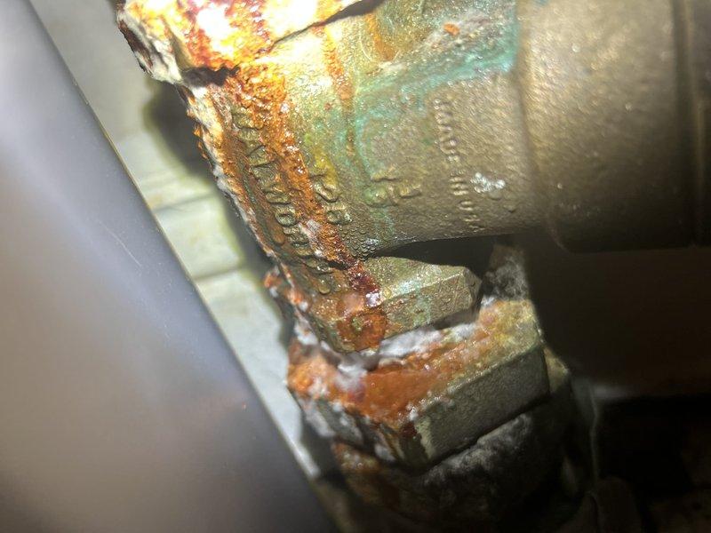 Responded to service call to provide estimate for emergency main water shut-off valve replacement located before the meter. Upon inspection, found existing shut-off valve exhibiting severe corrosion, rust buildup along brazed seams, and green copper oxidation indicating advanced deterioration and potential failure risk. Documented existing plumbing configuration including red-handled gate valve, white-handled ball valve, and associated piping assembly for estimate preparation.