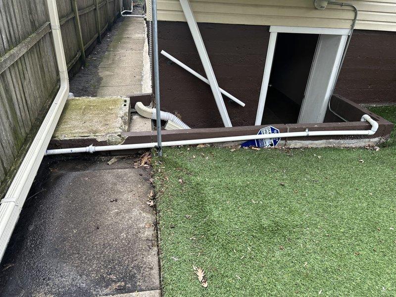 Responded to service call for malfunctioning sump pump system. Upon inspection, assessed sump pump installation including discharge piping configuration running from interior pit through exterior of residence. Verified PVC discharge line routing from basement/crawl space access point, through floor penetration, and out to exterior drainage termination point along side of building. Inspected pump chamber accessibility through floor-mounted access opening, confirmed proper pit construction and plumbing connections including vertical discharge pipe with appropriate fittings and floor flange assembly. Evaluated exterior discharge line installation including proper slope and termination away from foundation. System restored to proper working condition.