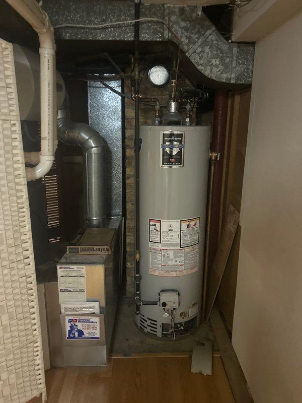 Responded to service call for residential water heater with no hot water production throughout home. Upon arrival, inspected 70-year-old home's utility closet containing tanked gas water heater unit. Performed diagnostic assessment of water heater system, examining gas control valve assembly, thermocouple connections, and pilot light operation. Checked gas supply line, brass fittings, and all copper piping connections for proper function. Evaluated temperature and pressure relief valve, inspected flue venting system, and verified proper combustion air supply. Tested electrical connections including thermocouple wiring and safety controls. Assessed overall unit condition including tank integrity, insulation, and manufacturer specifications label. Documented equipment configuration with utility room layout showing water heater positioned adjacent to HVAC furnace system with metal ductwork overhead. Restored hot water service to residence and verified proper operation of heating elements 