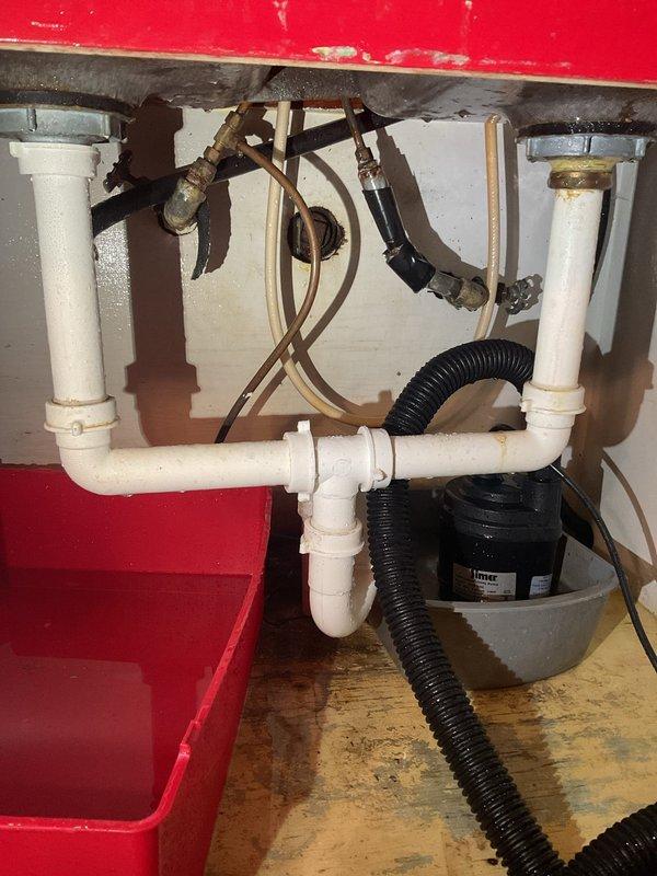 Conducted comprehensive drain and sewer assessment for commercial facility, focusing on under-sink plumbing infrastructure and main water supply systems. Documented existing P-trap configuration with white PVC piping, flexible supply lines, and pump assembly unit mounted beneath basin fixture, noting age-related discoloration on metal fittings and evidence of previous moisture exposure on surrounding wooden surfaces. Evaluated main water supply line with shutoff valve in utility area, identifying system conditions and photographing infrastructure for estimate preparation purposes.