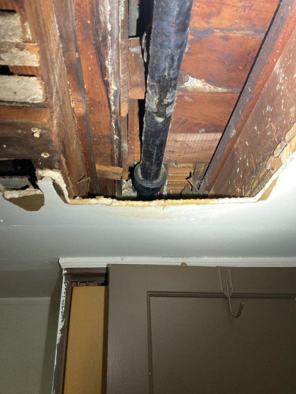 Responded to active water leak at bathroom sink drainage system in 110-year-old residential property. Upon inspection, identified leak source at junction where vertical and horizontal PVC drain pipes meet beneath sink, specifically at the P-trap connection point. Accessed plumbing through vanity cabinet and assessed compromised pipe joint assembly. Created ceiling access point to inspect vertical drain pipe routing through floor joists and verify extent of water damage to surrounding structural components. Documented existing white PVC P-trap configuration with elbow joint connections and vertical drain assembly. Photographed compromised plumbing connections and exposed ceiling cavity showing black drain pipe secured to wooden framing members. Completed four-hour service call addressing immediate leak concern and providing assessment of aging drainage infrastructure.