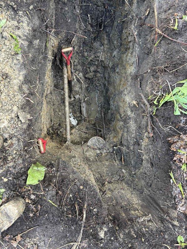 Excavated trench in saturated soil conditions to access underground plumbing system for repair work. Marked utility lines with red flagging tape for safety and reference, and prepared site for scheduled repair installation with proper drainage considerations due to high moisture content in work area.
