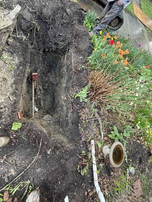 Excavated trench in saturated soil conditions to access underground plumbing system for repair work. Marked utility lines with red flagging tape for safety and reference, and prepared site for scheduled repair installation with proper drainage considerations due to high moisture content in work area.
