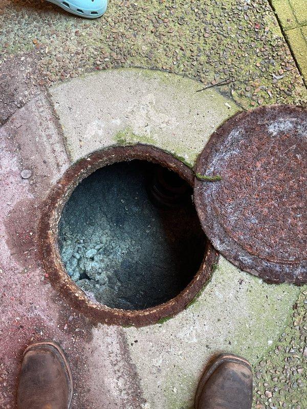 Responded to maintenance request for 12-unit condominium building requiring preventative sewer line rodding service. Upon arrival, located and accessed main sewer cleanout at ground level, which showed significant age and corrosion on access cover and rim assembly. Documented existing conditions including heavily rusted metal components, standing water with mineral deposits in access point, and deteriorated concrete surrounding the opening with biological growth and rust staining indicating long-term moisture exposure. Inspected interior sewer line access point revealing corroded pipe connections extending into main drainage system. Provided assessment of cleanout access conditions and sewer line maintenance requirements for the building's shared plumbing infrastructure.
