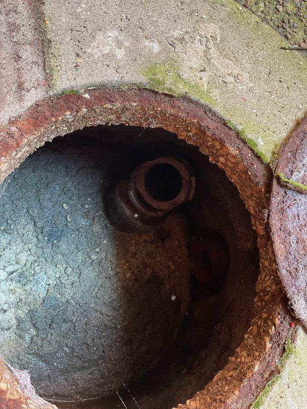 Responded to maintenance request for 12-unit condominium building requiring preventative sewer line rodding service. Upon arrival, located and accessed main sewer cleanout at ground level, which showed significant age and corrosion on access cover and rim assembly. Documented existing conditions including heavily rusted metal components, standing water with mineral deposits in access point, and deteriorated concrete surrounding the opening with biological growth and rust staining indicating long-term moisture exposure. Inspected interior sewer line access point revealing corroded pipe connections extending into main drainage system. Provided assessment of cleanout access conditions and sewer line maintenance requirements for the building's shared plumbing infrastructure.