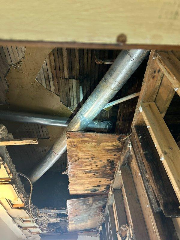 Responded to emergency call for burst pipe in ceiling discovered upon return from vacation. Upon arrival, found significant water damage with missing drywall exposing attic space and structural components. Inspection revealed burst pipe in ceiling cavity with extensive moisture damage to wooden joists and floor boards, evidenced by dark staining and deterioration throughout the affected area. Assessed water-damaged ductwork running through the space and evaluated extent of structural impact. Documented conditions showing prolonged water exposure affecting multiple wooden structural members and surrounding building materials. Performed thorough examination of plumbing system in affected area to identify source of pipe failure and assess necessary repairs to restore water integrity and prevent further damage.