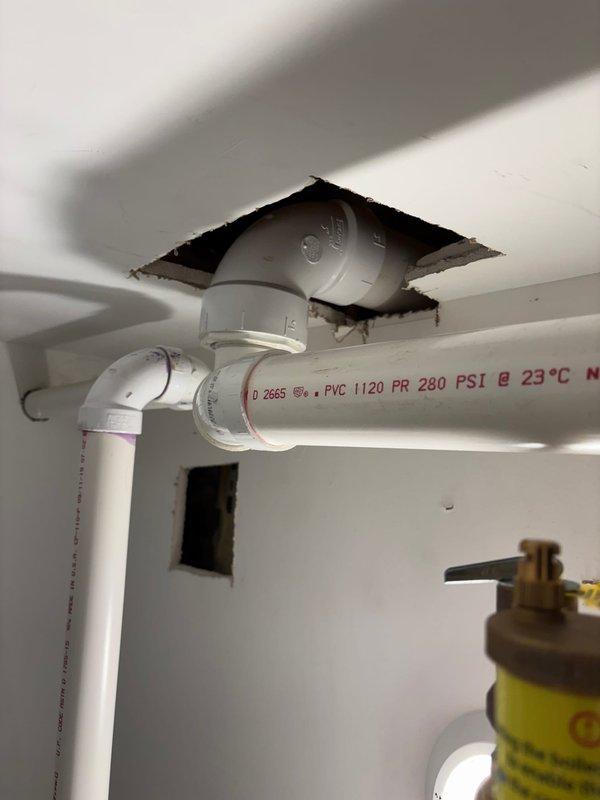 Responded to morning appointment request for plumbing repair assessment. Inspected under-sink plumbing configuration including white PVC drainage system with multiple fittings and connections, brass shut-off valve with pressure relief capabilities, and ceiling-mounted PVC supply lines rated at 280 PSI. Documented existing installation showing standard residential water supply and drainage components, ceiling access point with rough-cut opening for pipe routing, and overall system configuration for repair proposal.