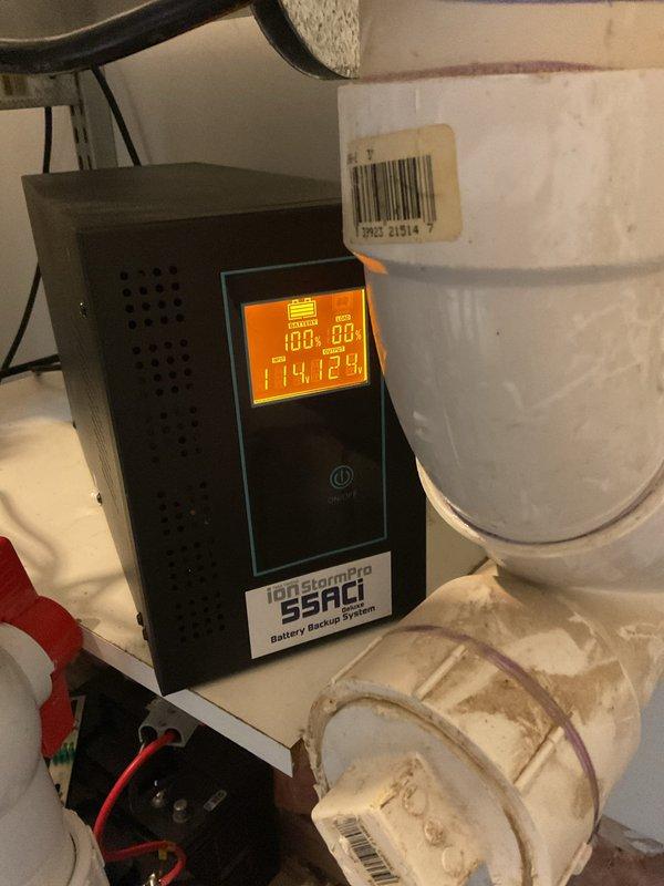 Installed and configured iOnstream SSACI battery backup system for sump pump operation. System was mounted in utility area with proper electrical connections established using appropriate gauge wiring. Battery backup unit displays operational voltage readings of 100.00V and 114.12V, confirming proper power supply and battery charge status. The backup system was integrated with existing sump pump installation featuring dual PVC discharge pipes and flexible rubber discharge hoses secured with stainless steel hose clamps. Verified all plumbing connections are watertight and functioning correctly, with active water management system operational and ready to provide continuous protection during power outages.