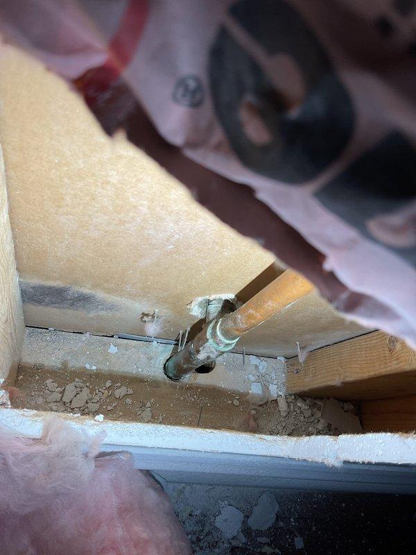 Responded to report of ceiling damage in living room located beneath bathroom and bedrooms in 19-year-old home. Upon arrival, inspected cracked ceiling with no active water dripping present. Cut access opening in ceiling to investigate suspected leak source above. Located corroded copper pipe joint showing signs of previous moisture intrusion and verdigris formation. Exposed PVC drain lines and wooden framing members were also inspected for damage. Documented conditions with photographs showing exposed piping, framing, and extent of ceiling damage requiring future drywall repair once plumbing work is completed.