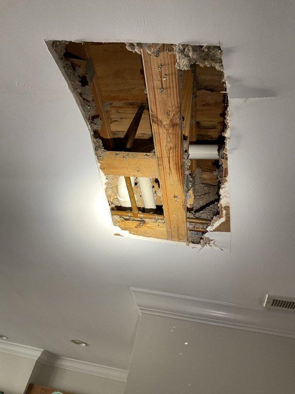 Responded to report of ceiling damage in living room located beneath bathroom and bedrooms in 19-year-old home. Upon arrival, inspected cracked ceiling with no active water dripping present. Cut access opening in ceiling to investigate suspected leak source above. Located corroded copper pipe joint showing signs of previous moisture intrusion and verdigris formation. Exposed PVC drain lines and wooden framing members were also inspected for damage. Documented conditions with photographs showing exposed piping, framing, and extent of ceiling damage requiring future drywall repair once plumbing work is completed.