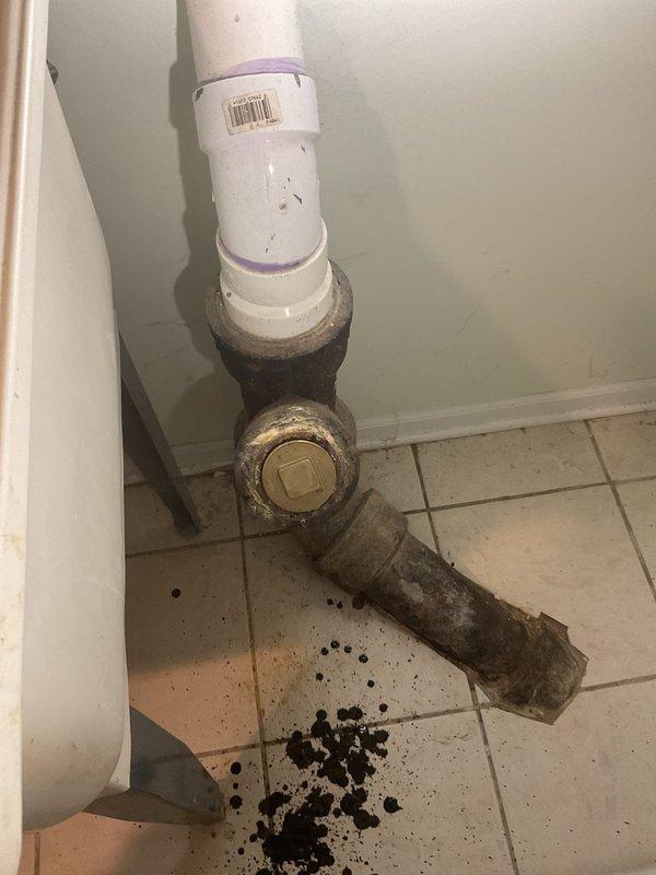 Responded to clogged drain service call at utility room location. Upon inspection, identified severely corroded horizontal drain pipe extending from brass fitting beneath utility sink. Pipe exhibited extensive deterioration with dark brown and black surface corrosion, with visible debris accumulation on floor tiles indicating drainage issues. Documented existing conditions including white PVC vertical drain line connection to corroded brass valve assembly. Return visit scheduled per management directive to complete necessary pipe replacement work. Additional technician assigned for Farrell fitting replacement with materials list to be coordinated prior to return service appointment.