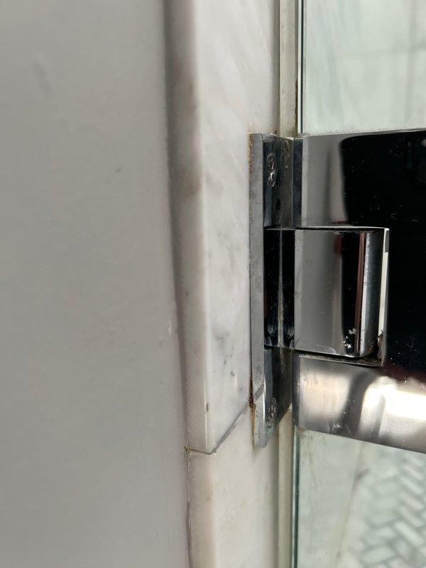 Inspected and assessed shower enclosure glass door assembly and surrounding fixtures. Examined chrome hinge mechanism connecting glass panel to door frame, noting normal wear patterns on metal components. Evaluated condition of white marble tile flooring with visible veining, identifying accumulated grime and discoloration along grout lines and corner seams. Assessed white door frame trim showing paint deterioration at floor junction. Documented decorative basket-weave pattern glass panel installation and mounting hardware functionality for maintenance records.
