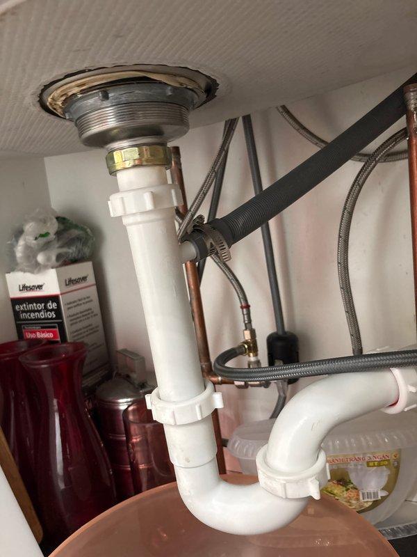 Responded to kitchen sink leak complaint. Upon inspection, identified active leak originating from P-trap assembly beneath sink basin. Observed water collection in bowl placed under plumbing, confirming persistent drip from drain connections. Examined white PVC P-trap configuration, chrome sink strainer, and associated threaded fittings for signs of deterioration or improper sealing. Documented existing plumbing setup including braided stainless steel supply lines, copper tubing, and dishwasher drain hose connection. Photographed leak source and overall drain assembly condition to assess repair requirements and provide customer with visual documentation of issue for approval to proceed with corrective work.