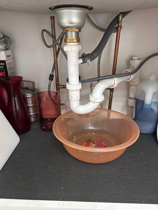 Responded to kitchen sink leak complaint. Upon inspection, identified active leak originating from P-trap assembly beneath sink basin. Observed water collection in bowl placed under plumbing, confirming persistent drip from drain connections. Examined white PVC P-trap configuration, chrome sink strainer, and associated threaded fittings for signs of deterioration or improper sealing. Documented existing plumbing setup including braided stainless steel supply lines, copper tubing, and dishwasher drain hose connection. Photographed leak source and overall drain assembly condition to assess repair requirements and provide customer with visual documentation of issue for approval to proceed with corrective work.