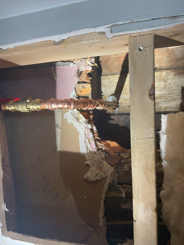 Responded to outdoor hose line leak in 121-year-old home where water supply had been shut off since January. Accessed plumbing system through crawl space/floor joist area and identified severely corroded copper pipe with compromised brass compression fittings as source of leak. Replaced deteriorated section of copper supply line and installed new brass shut-off valve with red handle. Secured new piping properly within existing floor joist cavity and verified all connections were leak-free before restoring water supply to outdoor spigot system.