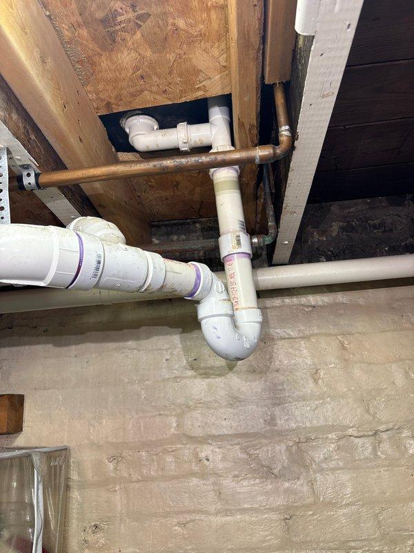Responded to water leak complaint originating from plumbing beneath bathtub. Accessed crawl space and inspected drain assembly, identifying active leak at PVC trap configuration. Found compromised joint connections with evidence of moisture damage on surrounding concrete floor. Evaluated both drainage system (white PVC with purple primer at joints) and adjacent copper water supply lines for integrity. Documented existing plumbing infrastructure including P-trap assembly, multiple PVC fittings, and copper piping showing age-related patina. Photographed affected area showing pipe routing through floor joists and subflooring for repair documentation and future reference.