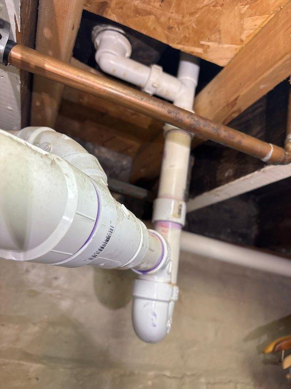 Responded to water leak complaint originating from plumbing beneath bathtub. Accessed crawl space and inspected drain assembly, identifying active leak at PVC trap configuration. Found compromised joint connections with evidence of moisture damage on surrounding concrete floor. Evaluated both drainage system (white PVC with purple primer at joints) and adjacent copper water supply lines for integrity. Documented existing plumbing infrastructure including P-trap assembly, multiple PVC fittings, and copper piping showing age-related patina. Photographed affected area showing pipe routing through floor joists and subflooring for repair documentation and future reference.