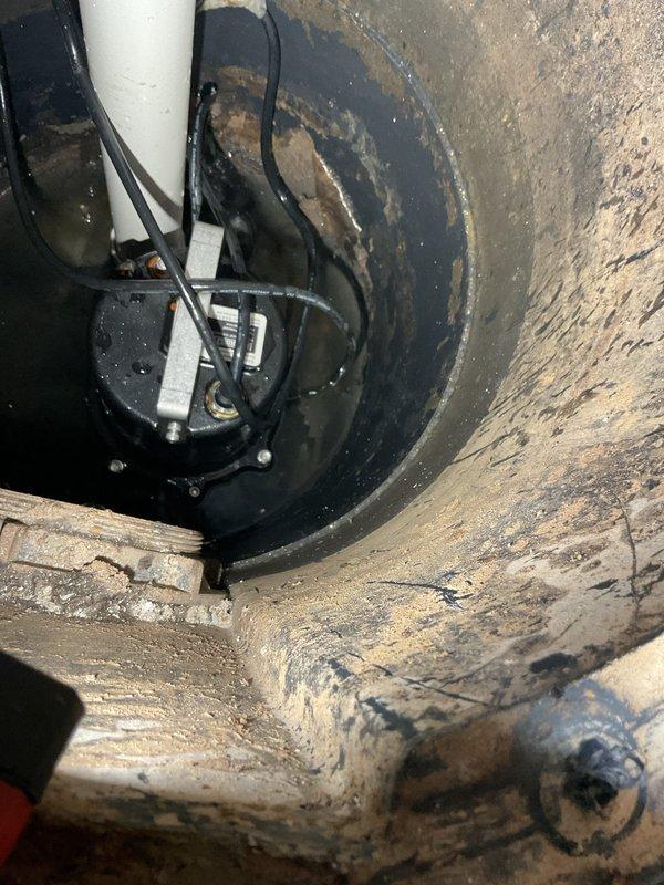 Responded to emergency call for ejector pump failure where another company had recently installed a new pump but water levels continued rising in the basin. Upon inspection, discovered the newly installed submersible ejector pump in the sump pit with water accumulating despite the replacement unit. Evaluated the complete waste pump system including the vertical PVC discharge piping, rubber coupling connections, and electrical connections to diagnose the ongoing water intrusion issue. Documented the condition of the installation, including the deteriorated wooden pit cover frame showing significant moisture damage, the discharge pipe assembly with multiple coupling sections, and the pump positioning within the basin to determine root cause of system malfunction and provide estimate for proper repairs.