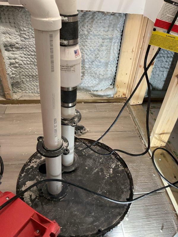 Responded to emergency call for ejector pump failure where another company had recently installed a new pump but water levels continued rising in the basin. Upon inspection, discovered the newly installed submersible ejector pump in the sump pit with water accumulating despite the replacement unit. Evaluated the complete waste pump system including the vertical PVC discharge piping, rubber coupling connections, and electrical connections to diagnose the ongoing water intrusion issue. Documented the condition of the installation, including the deteriorated wooden pit cover frame showing significant moisture damage, the discharge pipe assembly with multiple coupling sections, and the pump positioning within the basin to determine root cause of system malfunction and provide estimate for proper repairs.
