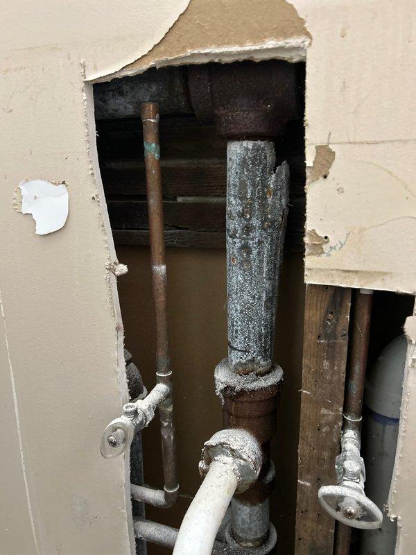 Accessed plumbing system through wall cavity and discovered extensive mold growth covering multiple pipes and interior surfaces. Removed contaminated section of drywall to expose affected plumbing assembly including vertical drain lines, copper supply pipes, and PVC drainage system. Addressed biological contamination on piping, fittings, and structural components within wall cavity. Inspected under-sink plumbing configuration with Charlotte-branded PVC pipes, metal hangers, and associated drain assemblies. Documented conditions of corroded pipe supports and multiple water supply and drain line connections for remediation planning.