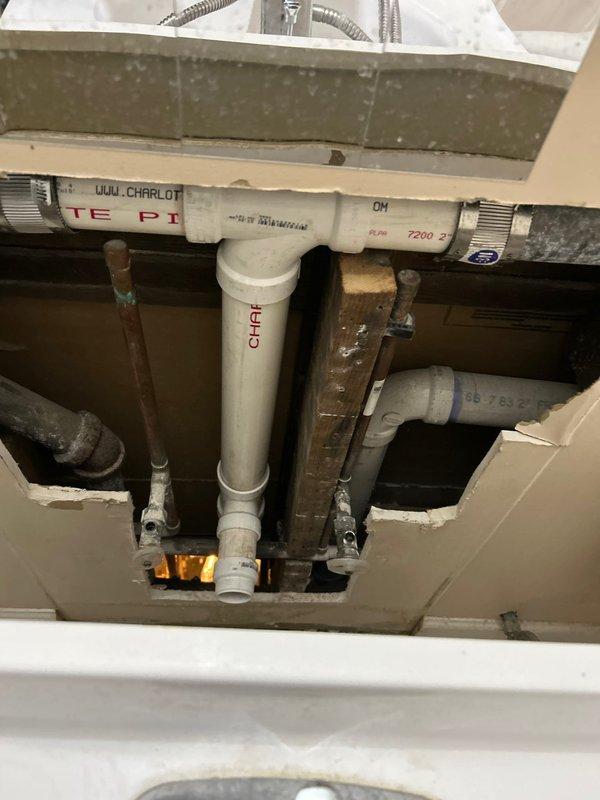 Accessed plumbing system through wall cavity and discovered extensive mold growth covering multiple pipes and interior surfaces. Removed contaminated section of drywall to expose affected plumbing assembly including vertical drain lines, copper supply pipes, and PVC drainage system. Addressed biological contamination on piping, fittings, and structural components within wall cavity. Inspected under-sink plumbing configuration with Charlotte-branded PVC pipes, metal hangers, and associated drain assemblies. Documented conditions of corroded pipe supports and multiple water supply and drain line connections for remediation planning.