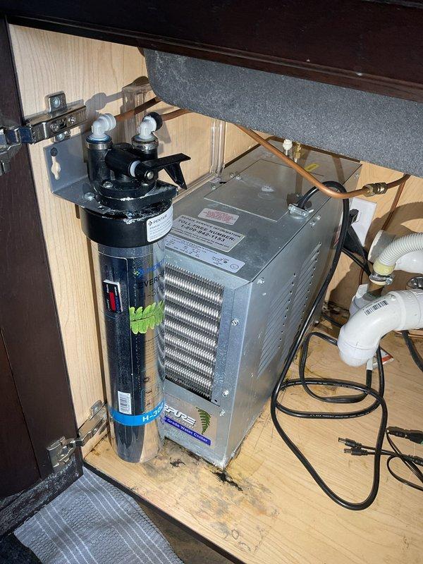 Responded to a leak at the connection point on an Everpure water filtration system installed in an under-cabinet location. Inspected the black cylindrical filter unit and associated plumbing connections, identifying the source of the leak at one of the top-mounted connection ports where white and gray supply lines interface with the filter housing. Tightened and resealed the compromised connection to eliminate the leak and restore proper system operation.