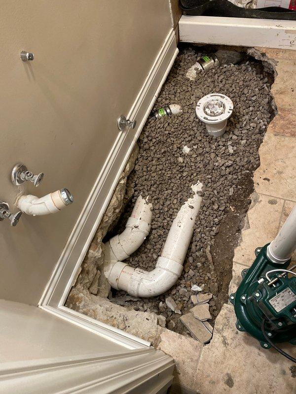 Completed main line drain and sewer installation in bathroom with full floor excavation to access and replace existing plumbing infrastructure. Installed new white PVC drain lines including P-trap configuration, straight drain pipes, and toilet flange assembly at proper elevations. Routed multiple diameter PVC drain and vent lines through existing wall framing from bathroom to basement level, securing pipes to wooden studs with appropriate hangers and supports. Applied purple primer and solvent cement to all PVC joints per code requirements. Installed sump pump system in bathroom floor cavity to manage water drainage. Prepared substrate with gravel aggregate base over concrete for proper drainage and pipe bedding. Coordinated plumbing rough-in with tub surround installation, positioning drain assemblies for compatibility with existing chrome fixtures and valves. Maintained proper slope on all horizontal drain runs to ensure adequate flow. All work performed to meet local plumbing codes