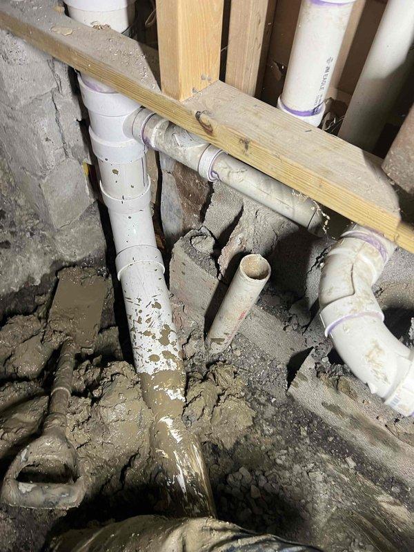 Completed main line drain and sewer installation in bathroom with full floor excavation to access and replace existing plumbing infrastructure. Installed new white PVC drain lines including P-trap configuration, straight drain pipes, and toilet flange assembly at proper elevations. Routed multiple diameter PVC drain and vent lines through existing wall framing from bathroom to basement level, securing pipes to wooden studs with appropriate hangers and supports. Applied purple primer and solvent cement to all PVC joints per code requirements. Installed sump pump system in bathroom floor cavity to manage water drainage. Prepared substrate with gravel aggregate base over concrete for proper drainage and pipe bedding. Coordinated plumbing rough-in with tub surround installation, positioning drain assemblies for compatibility with existing chrome fixtures and valves. Maintained proper slope on all horizontal drain runs to ensure adequate flow. All work performed to meet local plumbing codes