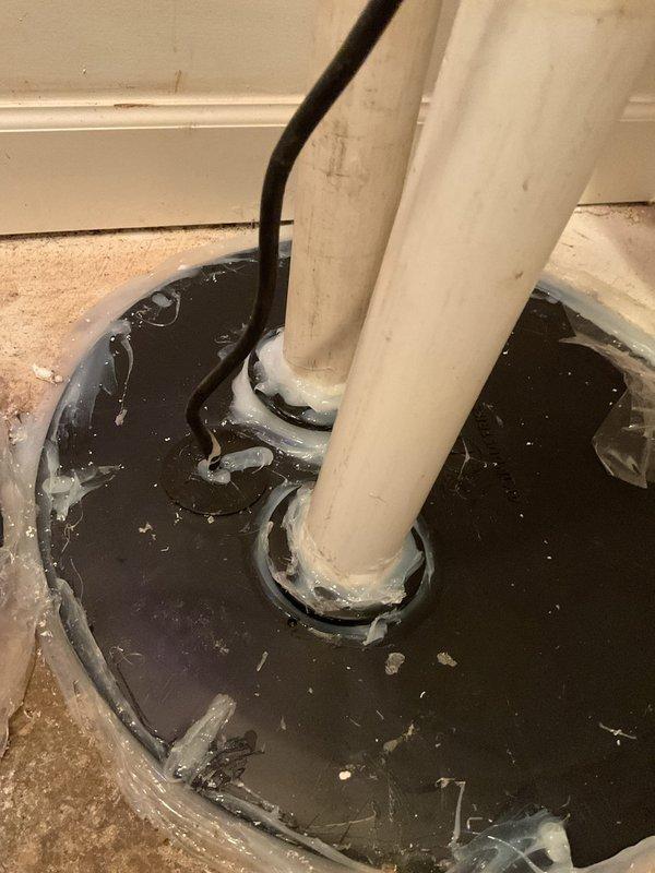 Responded to warranty callback for ejector pit installation completed six weeks prior. Upon inspection, confirmed customer complaint of sewer gas odor and drain fly infestation originating from improperly sealed ejector pit gaskets. Found rubber grommets around PVC discharge pipes compromised and deteriorated, allowing sewer gas escape into living space. Standing water observed in ejector pit with visible debris accumulation. Bathroom floor showed evidence of water overflow and unsanitary conditions with dark water pooling around toilet base. Removed failed rubber seals and cleaned pit opening perimeter. Installed new properly-sized rubber grommets around both discharge pipes, ensuring watertight seal to prevent gas infiltration. Verified all penetrations sealed and no gaps present around piping. Tested ejector pump operation and confirmed proper cycling. Cleaned surrounding area and removed standing water. Advised customer that drain fly issue should resolve within days as breeding so