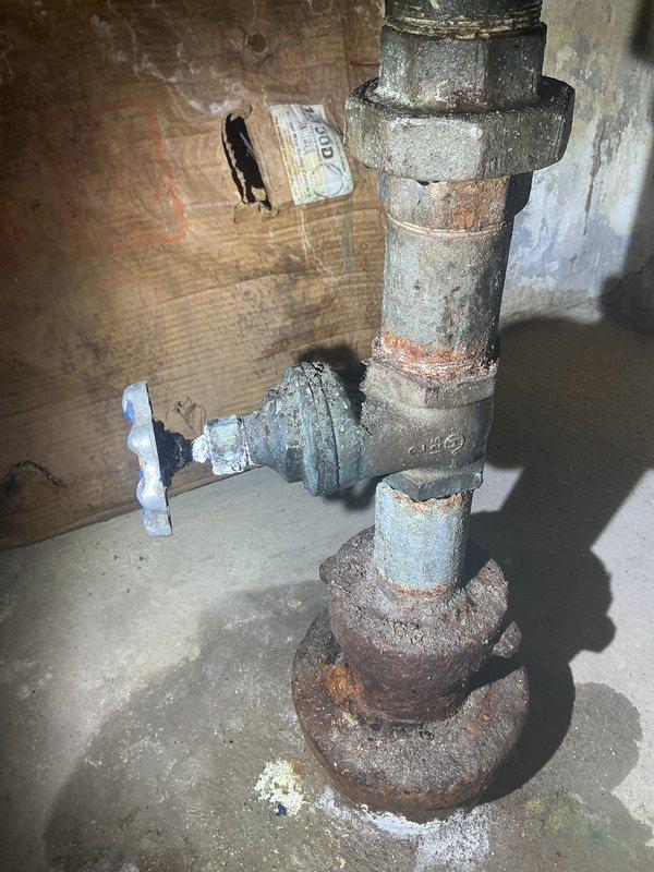 Conducted comprehensive assessment of aging water service line and associated plumbing infrastructure displaying severe corrosion and deterioration. Documented existing conditions of galvanized steel piping system including vertical riser, horizontal supply lines, gate valve assembly, and threaded fittings showing extensive rust, scaling, and compromised metal integrity. Identified moisture staining on concrete flooring near pipe base and evaluated structural condition of corroded connections requiring replacement. Photographed deteriorated plumbing components to support estimate preparation for complete water service line upgrade and valve replacement.