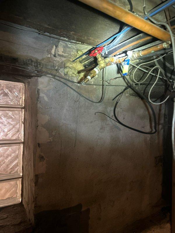 Responded to water leak affecting outdoor line beneath deck and garage area. Upon inspection, located compromised water supply line running through basement utility area with visible moisture damage on concrete wall surfaces and aging copper piping system. Assessed refrigerator water supply connection for modification as requested. Documented existing plumbing infrastructure including oxidized copper lines, shut-off valves, and routing configuration through exposed ceiling joists and along foundation walls.