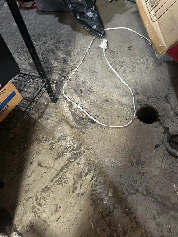 Inspected and assessed corroded cast iron drain piping beneath kitchen sink, documenting extensive rust and oxidation on vertical drain stack and P-trap assembly. Photographed existing plumbing configuration showing deteriorated metal pipes connected to white PVC components, and documented garage floor drain area with associated equipment for comprehensive repair planning.