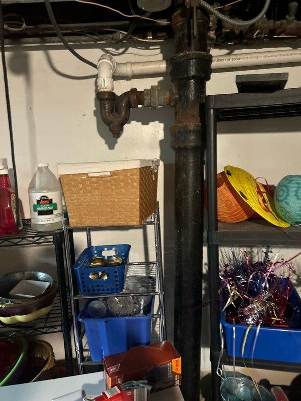 Inspected and assessed corroded cast iron drain piping beneath kitchen sink, documenting extensive rust and oxidation on vertical drain stack and P-trap assembly. Photographed existing plumbing configuration showing deteriorated metal pipes connected to white PVC components, and documented garage floor drain area with associated equipment for comprehensive repair planning.