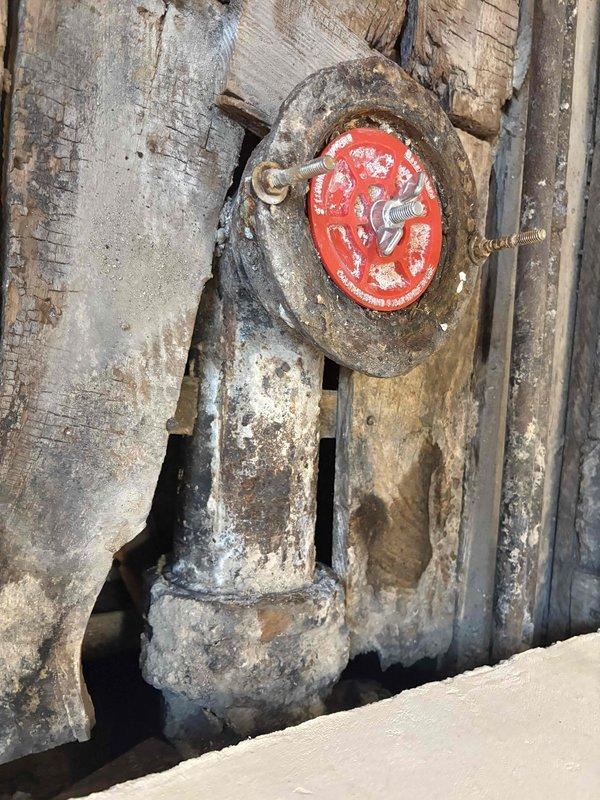 Conducted comprehensive assessment of deteriorated valve assembly and surrounding infrastructure for piping estimate. Documented extensively corroded gate valve with compromised red operating wheel showing significant paint failure and metal degradation. Evaluated aged wooden platform structure exhibiting severe moisture damage, structural separation between planks, and multiple areas of wood rot requiring replacement. Photographed existing piping connections and mounting points to determine scope of work for valve replacement and platform reconstruction necessary to restore system functionality and structural integrity.