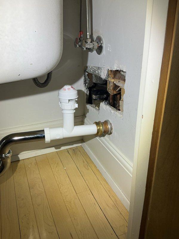 Responded to plumbing service call for kitchen sink drainage issues. Upon inspection, discovered water damage to cabinet interior wall and compromised plumbing connections beneath undermount stainless steel sink. Removed damaged drywall section to access deteriorated drain assembly and assess extent of leak damage. Replaced defective P-trap and drain fittings with new white PVC piping and chrome components. Installed proper compression fittings and ensured secure connections to prevent future leakage. Verified water heater supply lines and shutoff valve functionality in adjacent utility area. Tested all drain connections for proper flow and confirmed no active leaks present. Area prepared for drywall repair and restoration upon completion of plumbing work.
