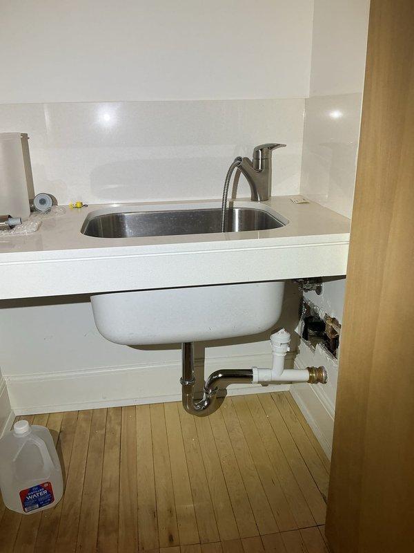 Responded to plumbing service call for kitchen sink drainage issues. Upon inspection, discovered water damage to cabinet interior wall and compromised plumbing connections beneath undermount stainless steel sink. Removed damaged drywall section to access deteriorated drain assembly and assess extent of leak damage. Replaced defective P-trap and drain fittings with new white PVC piping and chrome components. Installed proper compression fittings and ensured secure connections to prevent future leakage. Verified water heater supply lines and shutoff valve functionality in adjacent utility area. Tested all drain connections for proper flow and confirmed no active leaks present. Area prepared for drywall repair and restoration upon completion of plumbing work.
