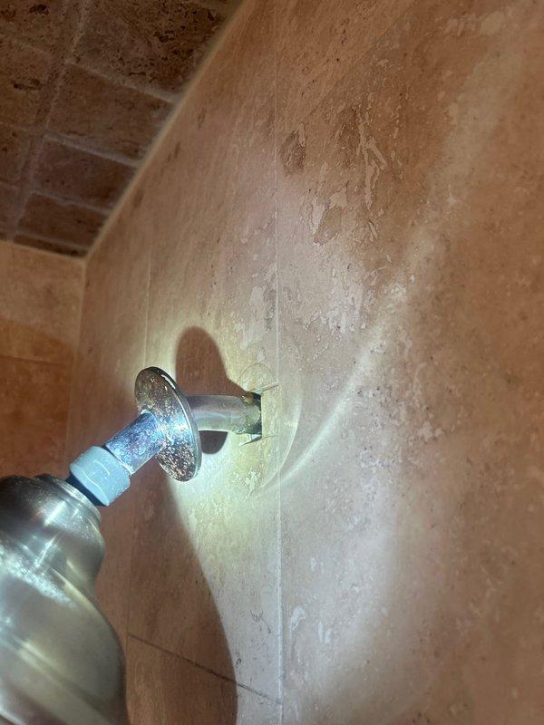 Responded to water leak complaint originating from ceiling below shower area during operation. Upon inspection of the shower enclosure, examined the chrome shower arm, handheld showerhead assembly, and associated wall penetrations through the travertine tile surround. Investigated potential leak sources including the shower arm escutcheon plate connection, showerhead mounting points, and the two chrome shower control valves. Assessed the integrity of grout lines and tile installations at wall corners and around all plumbing fixtures where water penetration could occur. Documented existing conditions of the shower assembly including the flexible hose connection with white plastic collar/gasket and all chrome fixtures for evidence of moisture intrusion or improper sealing that would allow water to bypass the shower pan and leak into the space below.