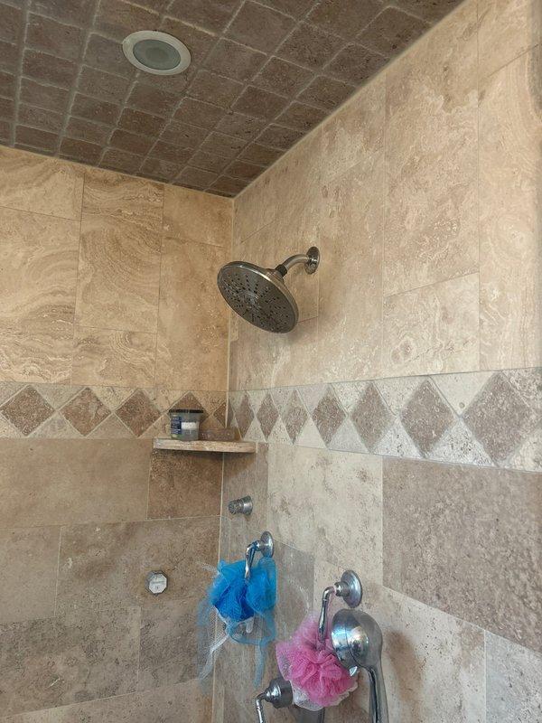 Responded to water leak complaint originating from ceiling below shower area during operation. Upon inspection of the shower enclosure, examined the chrome shower arm, handheld showerhead assembly, and associated wall penetrations through the travertine tile surround. Investigated potential leak sources including the shower arm escutcheon plate connection, showerhead mounting points, and the two chrome shower control valves. Assessed the integrity of grout lines and tile installations at wall corners and around all plumbing fixtures where water penetration could occur. Documented existing conditions of the shower assembly including the flexible hose connection with white plastic collar/gasket and all chrome fixtures for evidence of moisture intrusion or improper sealing that would allow water to bypass the shower pan and leak into the space below.