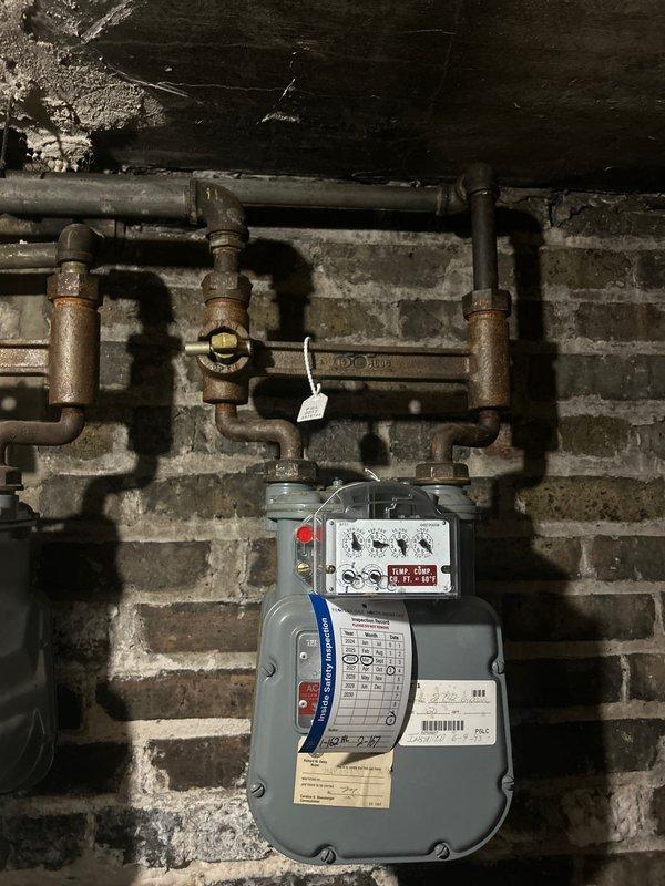 Completed assessment of second-floor gas piping system to provide re-pipe estimate. Inspected existing natural gas meter installation mounted on brick wall, noting significant corrosion and weathering on aged metal piping network, valves, and fittings. Evaluated utility room area containing laundry appliances to determine routing requirements and access points for new gas line installation. Documented current pipe configuration, valve locations, and connection points. Photographed existing conditions including corroded gas distribution piping and available workspace to develop accurate scope of work for complete second-floor gas line replacement project.