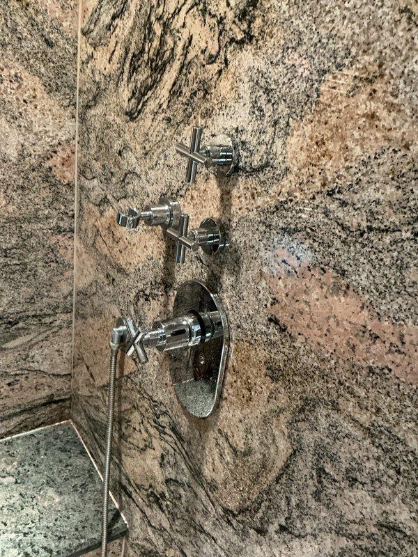 Completed installation of new chrome shower valve system in walk-in shower with polished granite wall panels. Installed three-piece fixture assembly including upper and middle cross-handle valve controls and lower main control unit with diverter valve and circular escutcheon plate, along with handheld shower head attachment. All fixtures tested and verified for proper water flow, temperature control, and diverter operation with no leaks detected.
