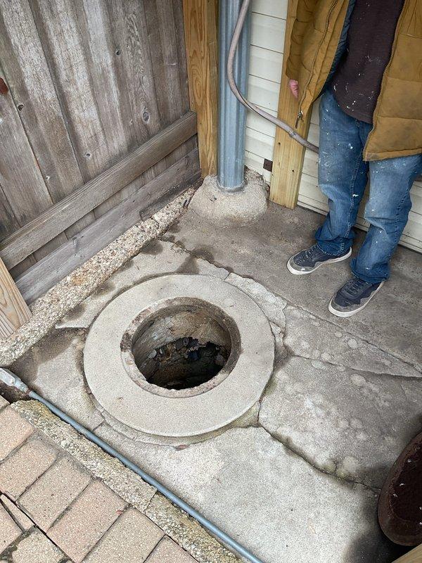 Responded to service call for catch basin maintenance at an 80-year-old three-flat residential property where drainage issues had persisted for several weeks. Upon arrival, conducted thorough inspection of backyard catch basin structure, documenting conditions through photographic evidence. 

Assessment revealed significant deterioration of the concrete catch basin, including extensive cracking throughout the interior walls, multiple areas of concrete spalling and delamination, and exposed substrate where surface material had broken away. The circular corrugated concrete pipe structure showed horizontal cracking patterns and structural degradation consistent with age-related weathering. Green discoloration and possible biological growth were observed on damaged interior sections, indicating prolonged moisture exposure. Standing water was present at the bottom of the basin with visible debris and particulate matter on the surface.

The concrete collar surrounding the basin opening at gr