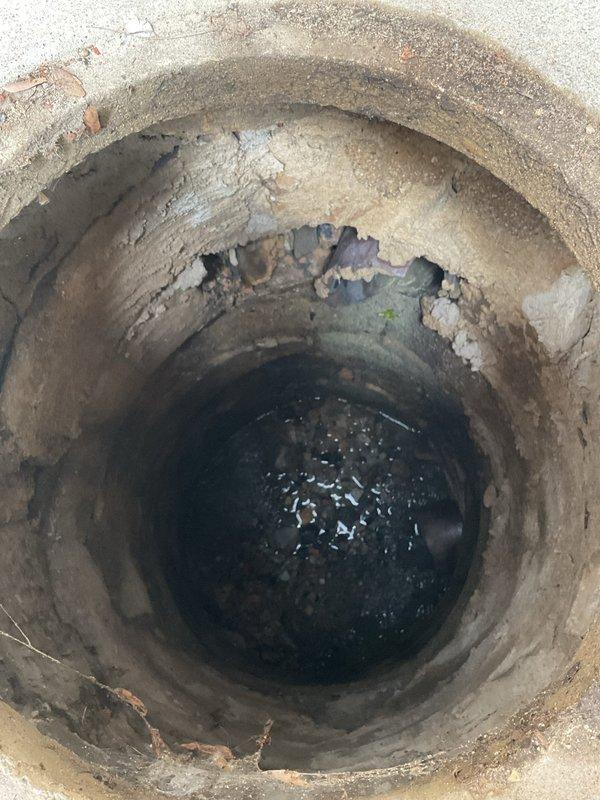 Responded to service call for catch basin maintenance at an 80-year-old three-flat residential property where drainage issues had persisted for several weeks. Upon arrival, conducted thorough inspection of backyard catch basin structure, documenting conditions through photographic evidence. 

Assessment revealed significant deterioration of the concrete catch basin, including extensive cracking throughout the interior walls, multiple areas of concrete spalling and delamination, and exposed substrate where surface material had broken away. The circular corrugated concrete pipe structure showed horizontal cracking patterns and structural degradation consistent with age-related weathering. Green discoloration and possible biological growth were observed on damaged interior sections, indicating prolonged moisture exposure. Standing water was present at the bottom of the basin with visible debris and particulate matter on the surface.

The concrete collar surrounding the basin opening at gr