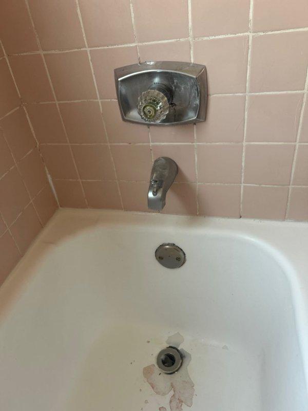 Responded to multiple plumbing concerns in a residential property including a toilet leaking at the base during flushing, potentially contributing to downstairs water damage, a shower temperature control valve malfunction preventing hot water delivery, and a deteriorated main water shut-off valve requiring replacement. Inspected vintage bathroom fixtures with pink tile surround, documenting the existing shower valve assembly with transparent faceplate and chrome tub spout showing age-related wear and mineral deposits. Assessed the toilet wax ring failure causing water seepage at the base and evaluated the corroded main shut-off valve that posed risk during operation.