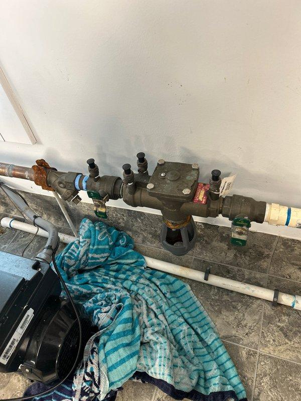 Responded to service call for dripping exterior valve requiring replacement. Upon inspection, identified backflow prevention device assembly with aged copper piping showing corrosion and deterioration, along with associated PVC connections and pressure regulation components. Documented existing plumbing configuration including flexible braided supply lines, brass fittings, and valve assembly mounted on interior wall above utility area flooring for estimate preparation and replacement recommendations.