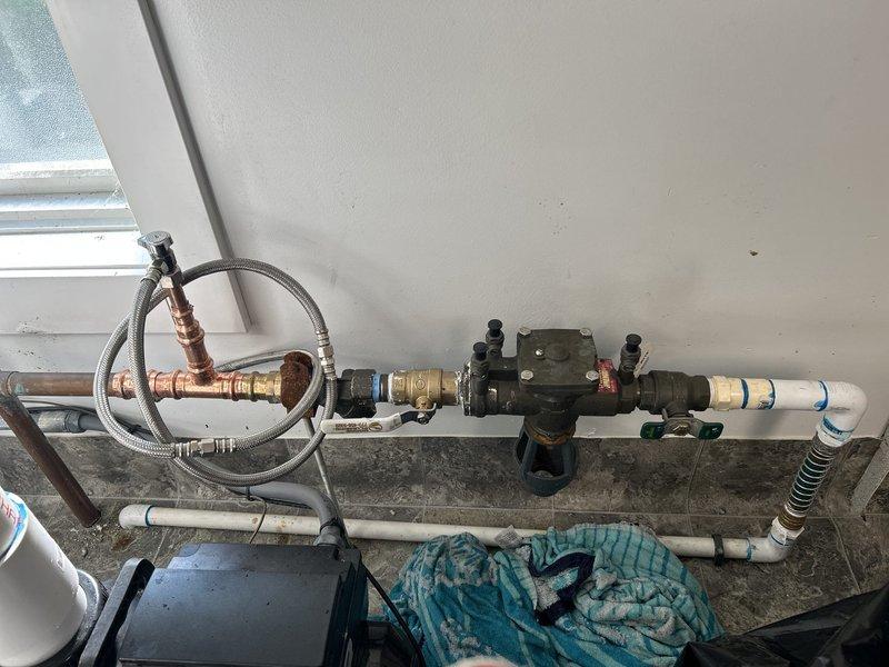 Responded to service call for dripping exterior valve requiring replacement. Upon inspection, identified backflow prevention device assembly with aged copper piping showing corrosion and deterioration, along with associated PVC connections and pressure regulation components. Documented existing plumbing configuration including flexible braided supply lines, brass fittings, and valve assembly mounted on interior wall above utility area flooring for estimate preparation and replacement recommendations.
