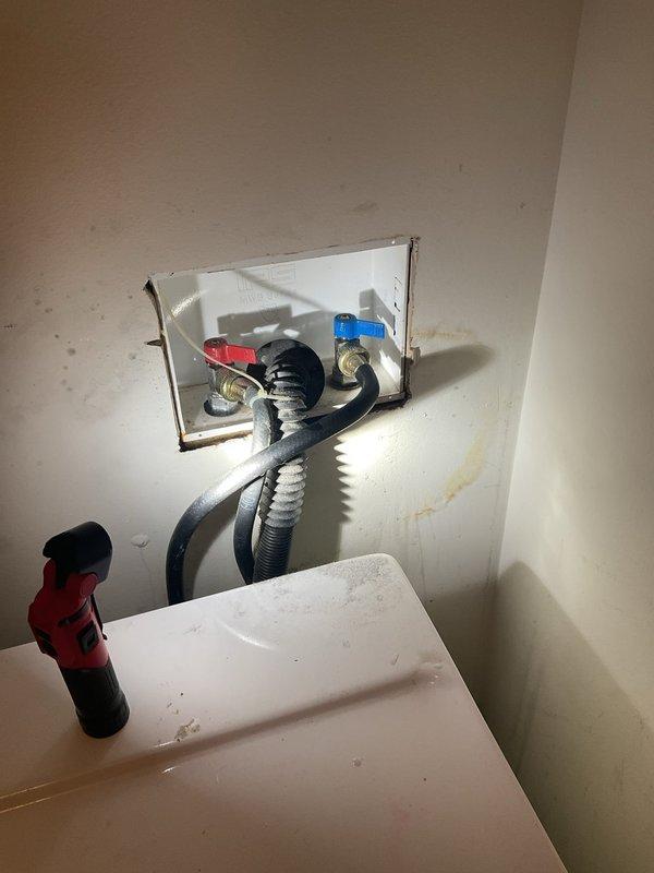 Conducted commercial drain and sewer estimate involving inspection of under-sink plumbing assembly with wall access panel. Assessment included evaluation of flexible supply lines, corrugated drainage tubing, shut-off valves with blue handles, and brass fittings for potential replacement or repair needs. Documented existing water staining on wall surfaces and multi-port valve connections requiring attention.