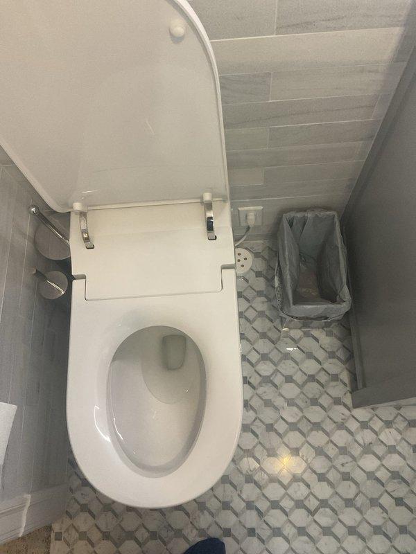 Responded to request for estimate to reposition toilet approximately 2 inches to center it properly in bathroom. Assessed existing toilet installation, floor tile configuration, and plumbing rough-in location to determine feasibility of relocation. Documented current toilet placement and surrounding bathroom layout including pebble-pattern floor tile and wall-mounted fixtures for estimate preparation.